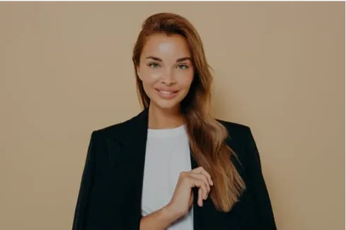 Portrait d'une femme aux longs cheveux bruns portant un blazer noir sur un t-shirt blanc, souriant avec un fond neutre.