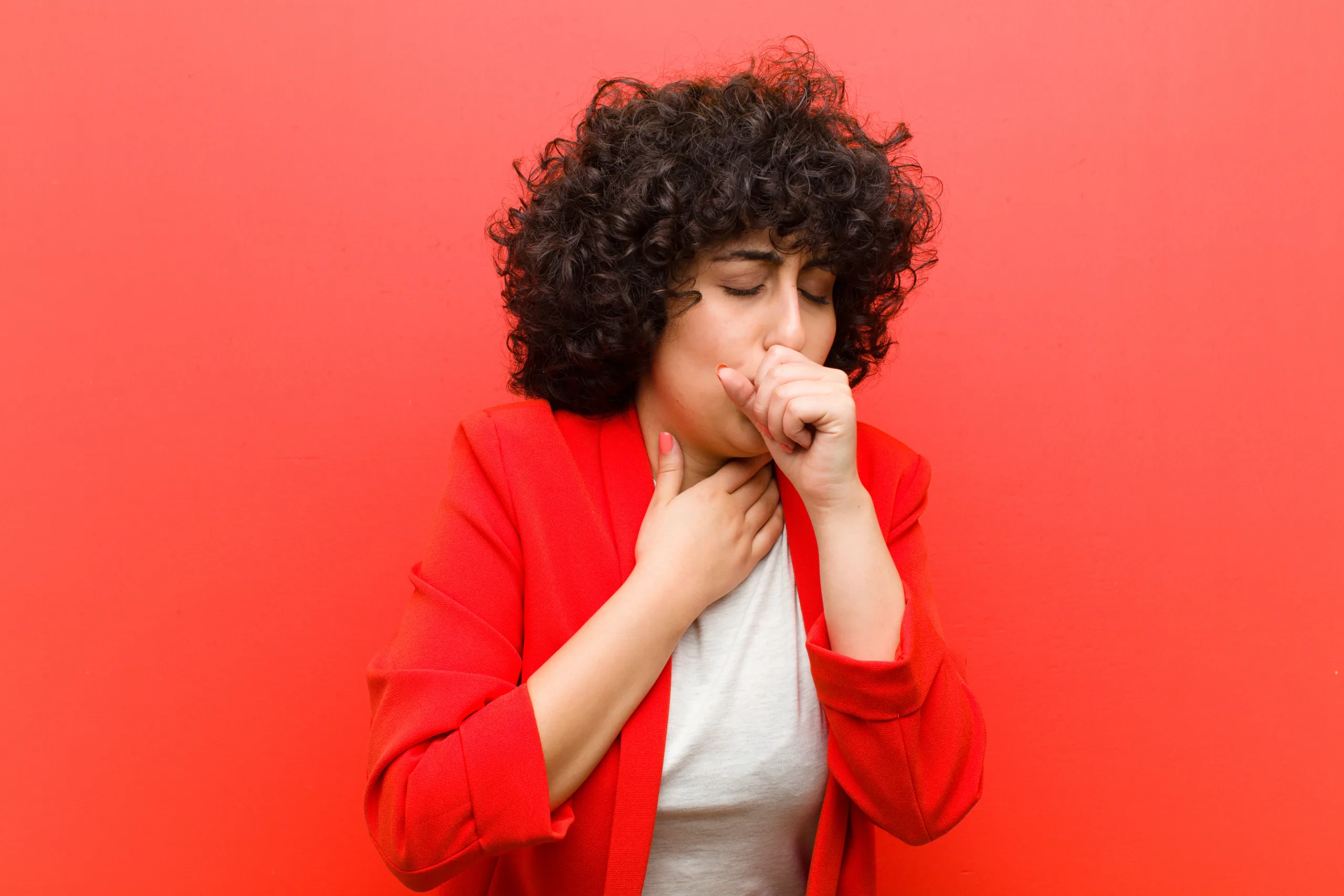 feeling ill with a sore throat and flu symptoms, coughing with mouth covered Jeune femme aux cheveux bouclés, portant un blazer rouge, se tenant la gorge et toussant, semblant souffrir d'un mal de gorge, devant un mur rouge vif.